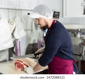 Young bearded man wearing red apron and mesh cap with visor, professional butcher, cutting slab of beef with large knife in processing room of supermarket, preparing meat products for sale - Powered by Shutterstock