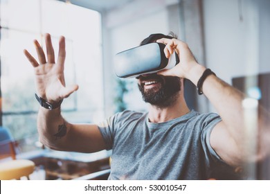 Young bearded man wearing virtual reality glasses in modern interior design coworking studio. Smartphone using with VR goggles headset. Horizontal,flares effect, blurred background