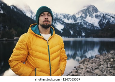 Young bearded man wearing yellow puff jacket and green hat standing near a mountain lake in winter. Snow mountain on background.  