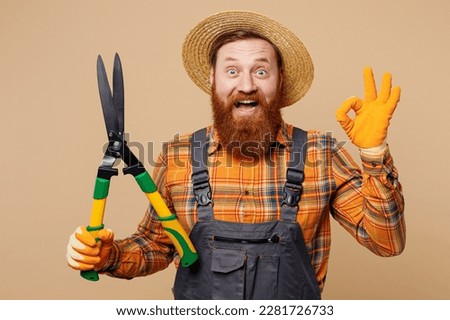 Young bearded man wears straw hat overalls work in garden hold in hand hedge shears scissorries show ok okay isolated on plain pastel light beige color background studio portrait. Plant caring concept