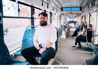 Young bearded man in white shirt and wristwatch sitting on passenger seat of modern train and looking at camera at daytime - Powered by Shutterstock