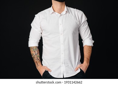 A Young Bearded Man In A White Shirt And Tattoos Stands On A Black Background.