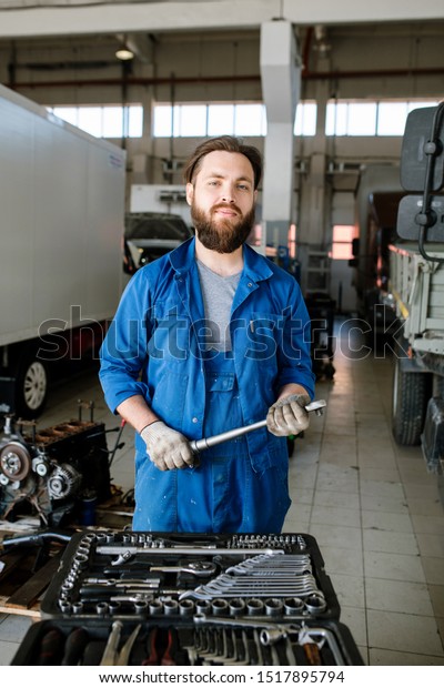 Young bearded\
master of technical repair standing inside workshop in front of\
camera with lorries on\
background
