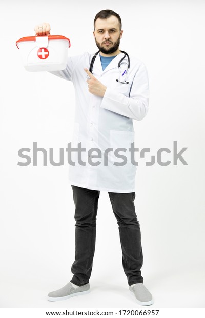 young bearded paramedic holding\
red cross medical kit pointing with index finger full height\
portrait isolated on white background copy space. emergency\
concept