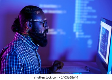 Young Bearded Programmer In Eyeglasses Looking At Computer Monitor And Examining Software In Dark Office