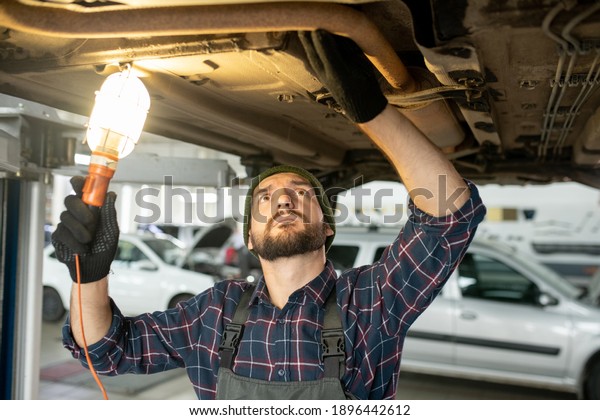 Young bearded repairman in gloves and uniform\
holding lamp while checking loosen details of underneath part of\
the car before repair