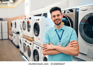 A Young Bearded Sales Assistant In Uniform Standing With His Arms Crossed In A Home Appliances And Electronics Store Against The Backdrop Of Washing Machines.