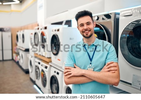 A young bearded sales assistant in uniform standing with his arms crossed in a home appliances and electronics store against the backdrop of washing machines.