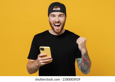 Young bearded tattooed man 20s he wears casual black t-shirt cap hold in hand use mobile cell phone do winner gesture isolated on plain yellow wall background studio portrait. Tattoo translate fun.