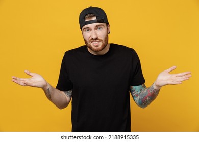 Young Bearded Tattooed Man 20s He Wears Casual Black T-shirt Cap Spread Hands Shrugging Shoulders Looking Puzzled, Have No Idea Isolated On Plain Yellow Wall Background Studio. Tattoo Translate Fun