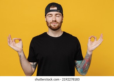 Young Bearded Tattooed Man 20s He Wears Casual Black T-shirt Cap Hold Spread Hands In Yoga Om Aum Gesture Relax Meditate Try To Calm Down Isolated On Plain Yellow Wall Background. Tattoo Translate Fun