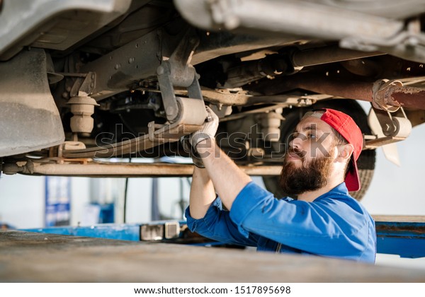 Young bearded\
technician of machine repair service fixing detail while standing\
under broker car inside\
workshop