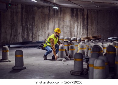Young bearded worker in vest with helmet on head trying to lift heavy concrete pillar while crouching in tunnel in construction process.