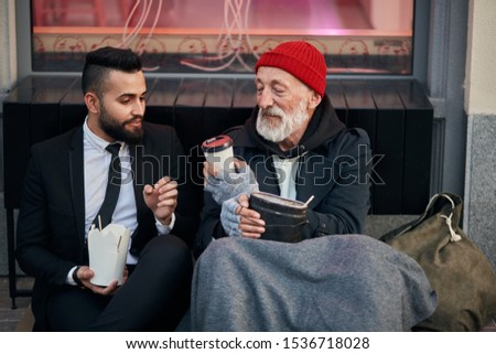 Young beardy man in suit sitting with beggar on floor on street and give cup of coffee. Different segments of society, social inequality