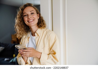 Young beatiful attractive smiling happy curly woman holding phone and looking at camera while standing at home - Shutterstock ID 2202148837