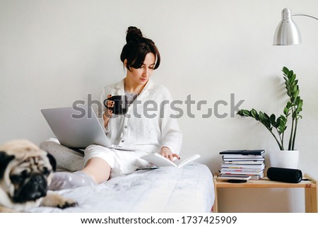 Young beatiful girl is reading a book and drinks tea on bed near a dog at home in isolation. Home office. Concept of working and studying in isolation.