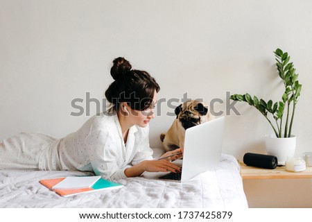 Young beatiful girl is working with laptop on bed near a dog at home in isolation. Home office. Concept of working and studying in isolation.