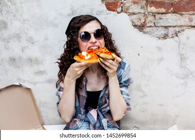 young beaty girl eat street food pizza and drink coffee at the city against the wall. teenager hold slice italy snack. woman wearing plaid shirt, jeans and black hat