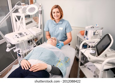 Young beautician sitting beside female client. Young woman with cosmetic mask on her face lying on daybed during skincare procedure in beauty salon. Concept of skincare, beauty and wellness.