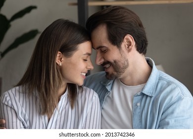 Young beautiful 30s loving married couple sit on couch enjoy romantic relation. Happy spouses spend time together at home feeling love, touch foreheads enjoy moment of tenderness, caress, date concept