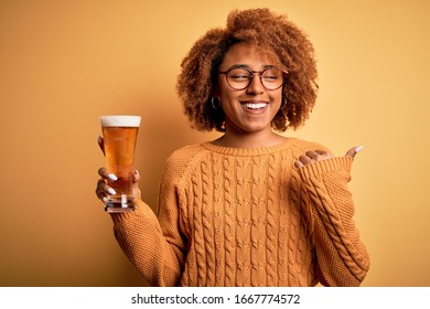Young beautiful African American afro woman with curly hair drinking glass of beer pointing and showing with thumb up to the side with happy face smiling