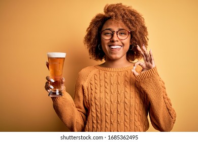 Young beautiful African American afro woman with curly hair drinking glass of beer doing ok sign with fingers, excellent symbol