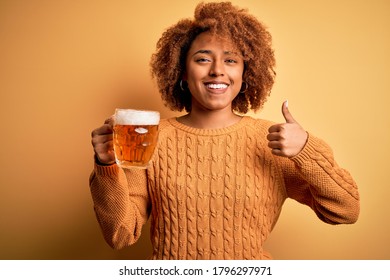 Young beautiful African American afro woman with curly hair drinking jar of beer happy with big smile doing ok sign, thumb up with fingers, excellent sign