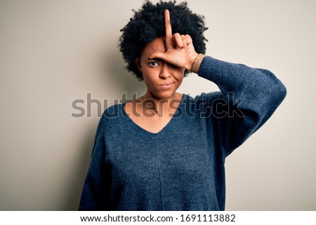 Young beautiful African American afro woman with curly hair wearing casual sweater making fun of people with fingers on forehead doing loser gesture mocking and insulting.