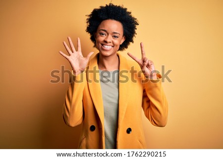 Young beautiful African American afro businesswoman with curly hair wearing yellow jacket showing and pointing up with fingers number seven while smiling confident and happy.