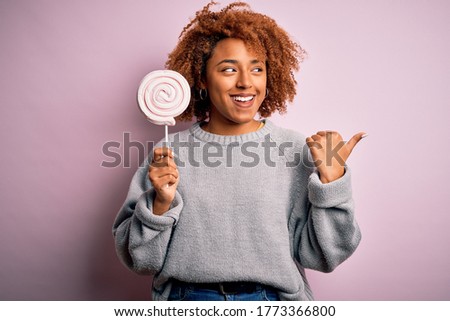 Young beautiful African American afro woman with curly hair eating sweet candy pointing and showing with thumb up to the side with happy face smiling