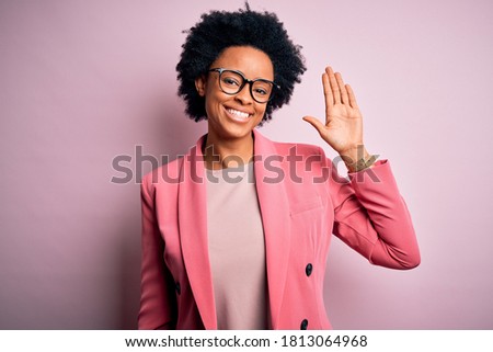 Young beautiful African American afro businesswoman with curly hair wearing pink jacket Waiving saying hello happy and smiling, friendly welcome gesture
