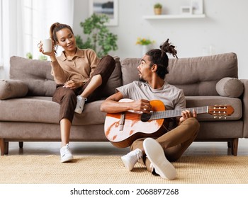 Young beautiful african american family couple playing acoustic guitar at home while relaxing together in living room during weekend, young man plays on musicial instrument for happy woman on sofa - Powered by Shutterstock