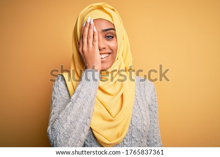 Young beautiful african american girl wearing muslim hijab over isolated yellow background covering one eye with hand, confident smile on face and surprise emotion.