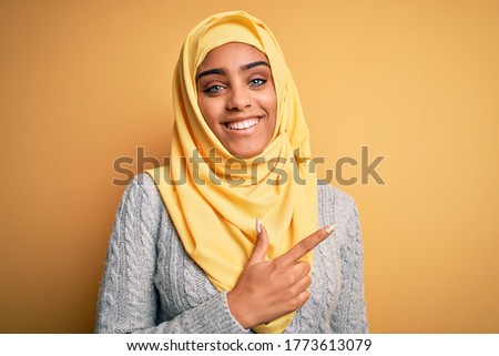 Young beautiful african american girl wearing muslim hijab over isolated yellow background cheerful with a smile on face pointing with hand and finger up to the side with happy and natural expression