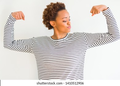 Young beautiful african american woman wearing stripes sweater over white background showing arms muscles smiling proud. Fitness concept.