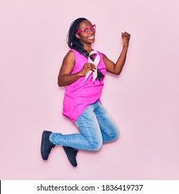 Young Beautiful African American Woman Wearing Glasses Smiling Happy. Jumping With Smile On Face Holding Pink Cancer Ribbon Celebrating With Fist Up Over Isolated Background