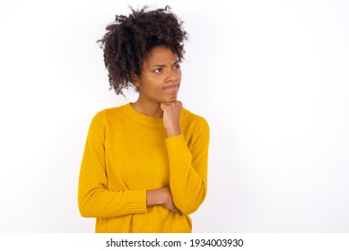 young beautiful African American woman wearing yellow sweater against white wall with hand under chin and looking sideways with doubtful and skeptical expression, suspect and doubt. - Powered by Shutterstock