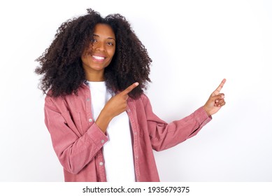 young beautiful African American woman wearing pink jacket against white wall indicating finger empty space showing best low prices, looking at the camera