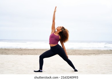 Young beautiful African American woman in work out clothing exercises at the beach doing the warrior pose with one arm up                               