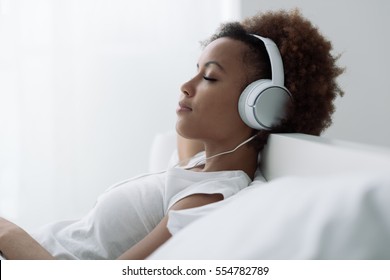 Young Beautiful African American Woman Relaxing And Listening To Music Using Headphones, She Is Lying In Bed