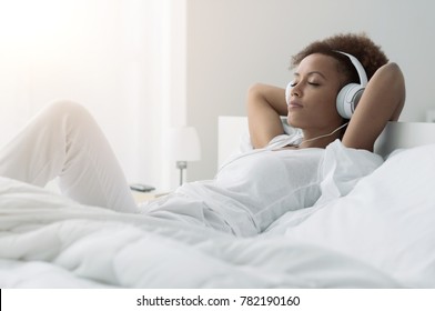 Young Beautiful African American Woman Relaxing And Listening To Music Using Headphones, She Is Lying In Bed