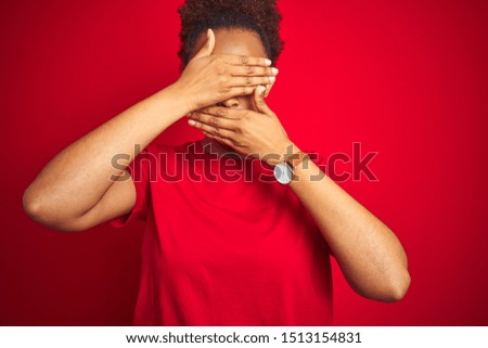 Young beautiful african american woman with afro hair over isolated red background Covering eyes and mouth with hands, surprised and shocked. Hiding emotion