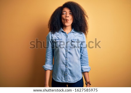 Young beautiful african american woman with afro hair standing over yellow isolated background making fish face with lips, crazy and comical gesture. Funny expression.