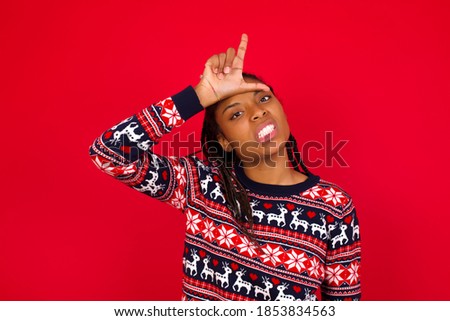 Young beautiful African American woman wearing Christmas sweater, against red background making fun of people with fingers on forehead doing loser gesture mocking and insulting.