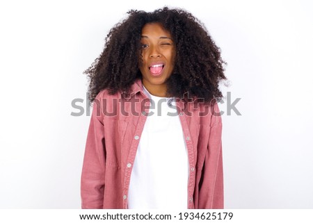 young beautiful African American woman wearing pink jacket against white wall sticking tongue out happy with funny expression. Emotion concept.