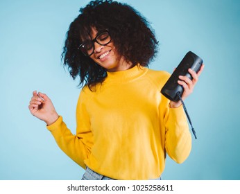 Young Beautiful African Woman Enjoying And Dancing At Blue Background. Modern Trendy Girl With Afro Hairstyle Listening To Music By Wireless Portable Speaker.