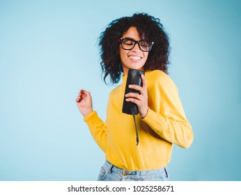 Young Beautiful African Woman Enjoying And Dancing At Blue Background. Modern Trendy Girl With Afro Hairstyle Listening To Music By Wireless Portable Speaker.