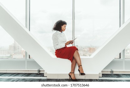 Young beautiful African-American businesswoman with a tablet PC is sitting on a load-bearing beam of a modern bright business office passageway next to a panoramic window with urban skyline outside