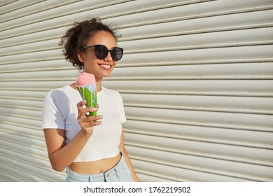 a young beautiful African-American girl in a white t-shirt and light jeans eats ice cream against a light wall on a Sunny day. - Powered by Shutterstock