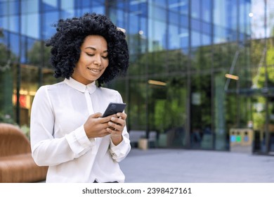 Young beautiful African-American woman walking street outside office building, businesswoman holding a phone i hands, smiling contentedly, browsinginternet and using an application on a smartphone.